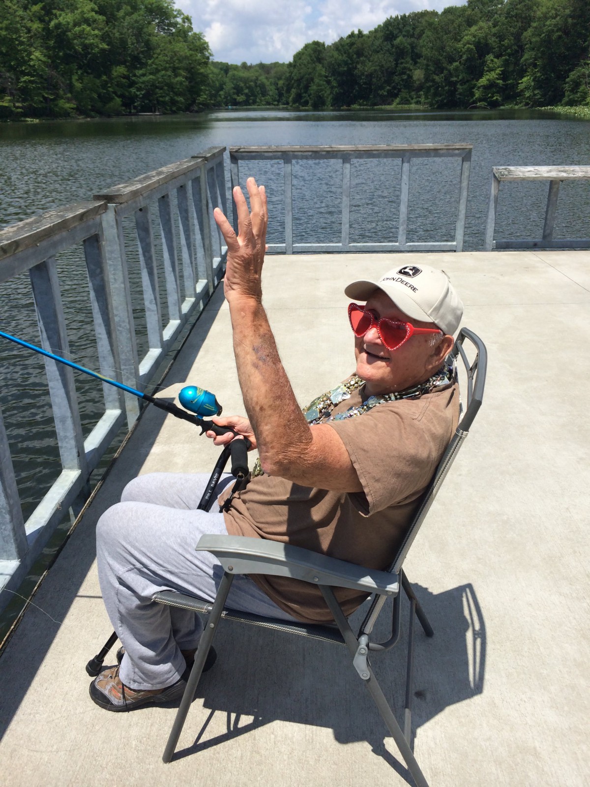 A man enjoying a sunny day fishing on a pier by the lake, wearing red sunglasses and a hat.
