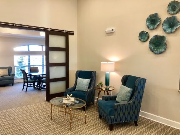 A cozy living room nook with two blue armchairs, a round glass coffee table, and decorative wall art.