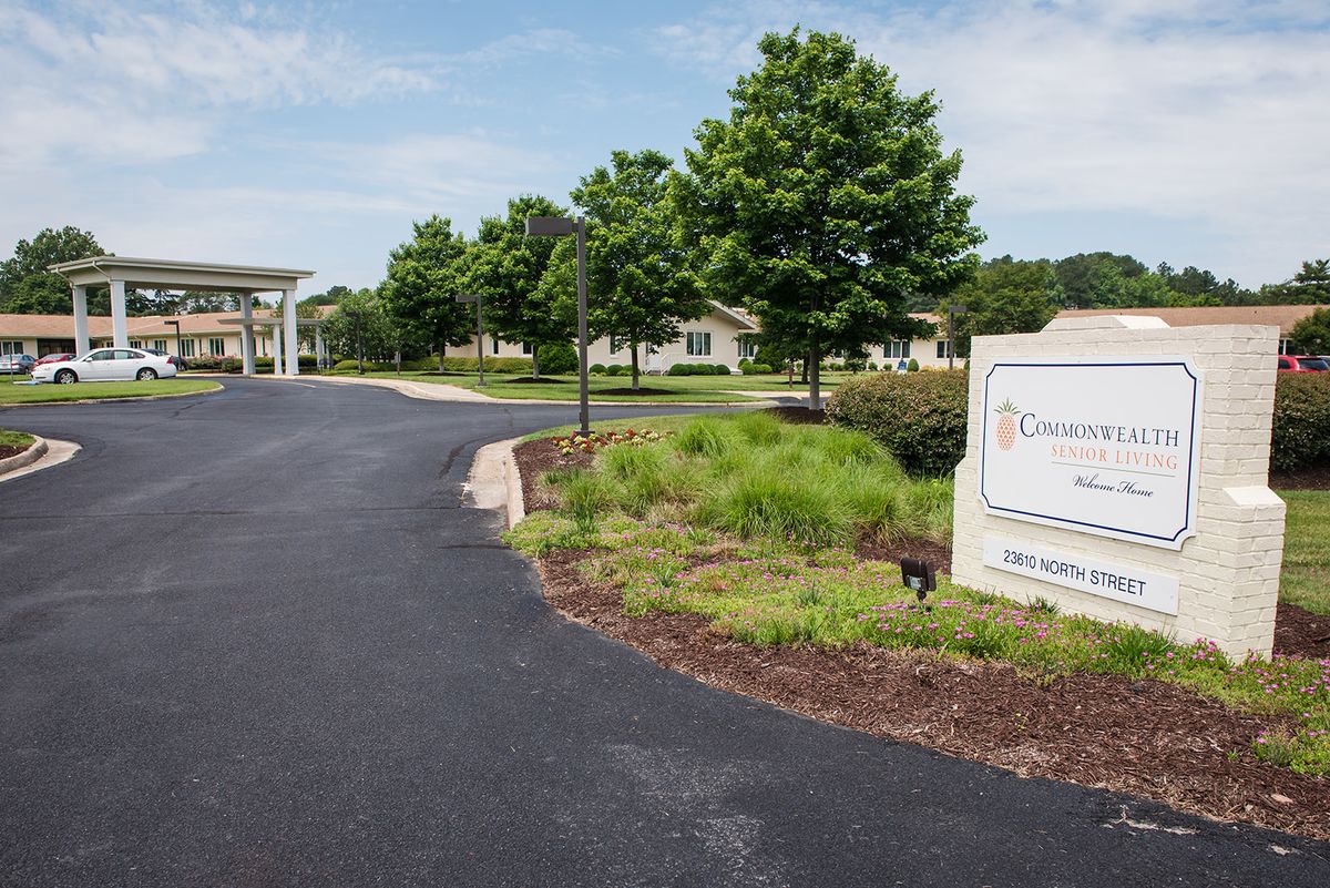 A paved road leading to a building, surrounded by trees and landscaped greenery, with a sign on the right.