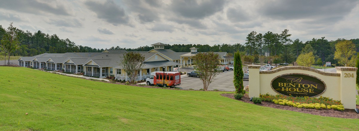 A building with a parking lot and a red bus parked outside, set against a backdrop of trees and a cloudy sky.