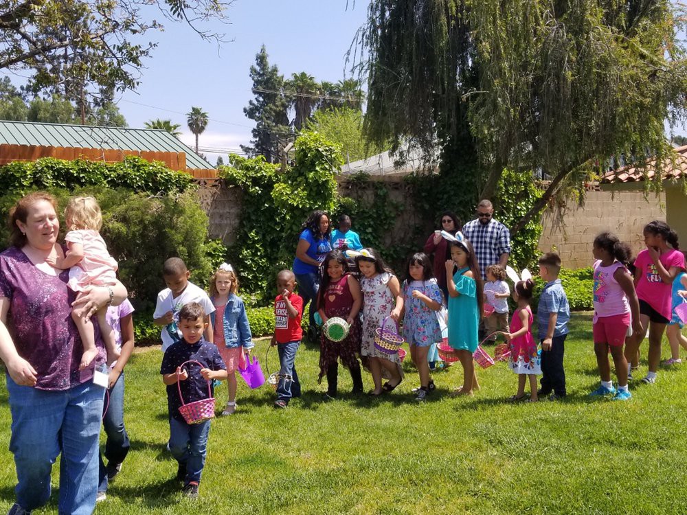 A group of children and adults are gathered outdoors in a garden setting, enjoying a sunny day.