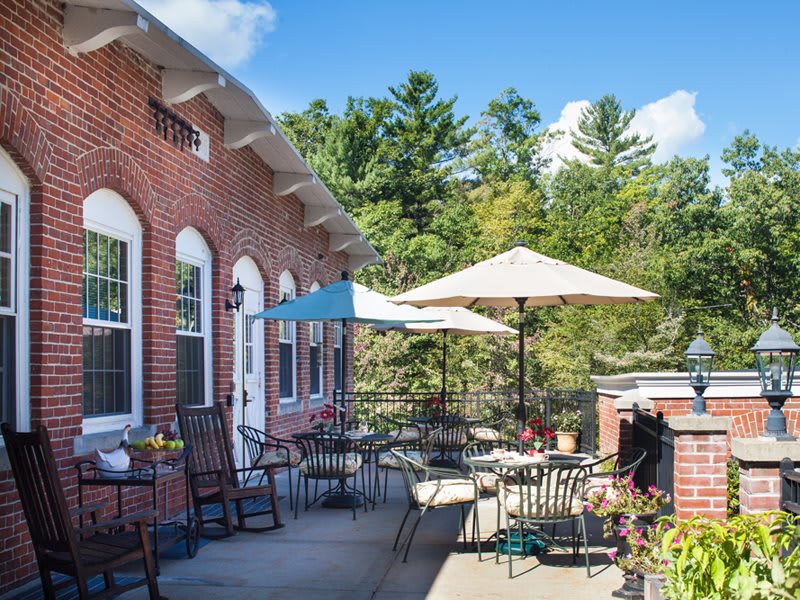 A cozy outdoor patio with tables, chairs, and umbrellas adjacent to a brick building, surrounded by trees under a clear blue sky.