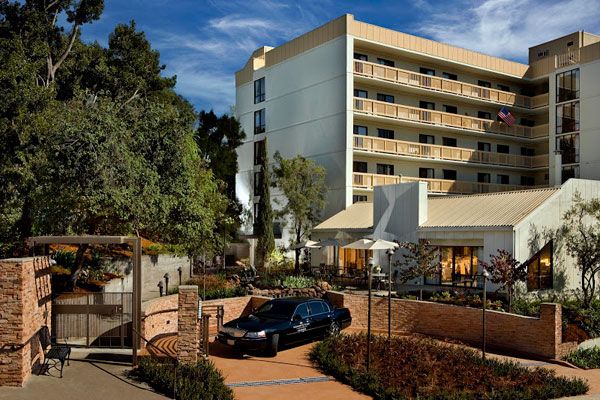 A modern hotel building with a parked car in front, surrounded by trees and landscaping under a sunny sky.