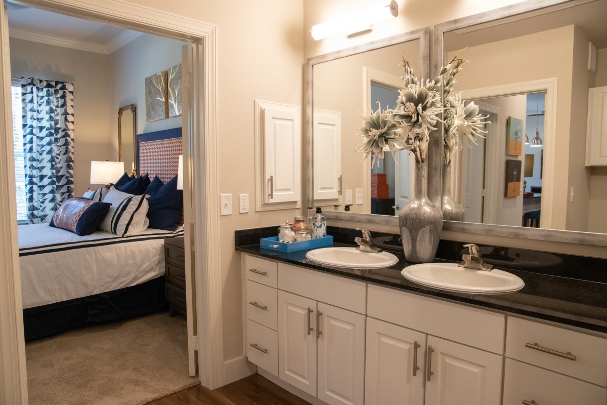A modern bathroom with double sinks and mirrors, featuring a decorative vase with flowers and a view into the adjoining bedroom.