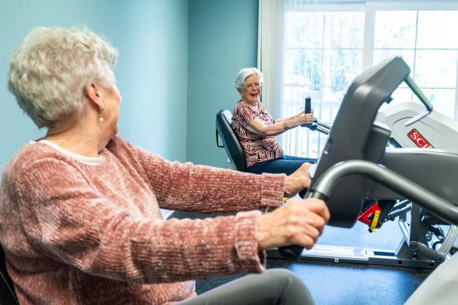 Two elderly women exercising on gym machines indoors.
