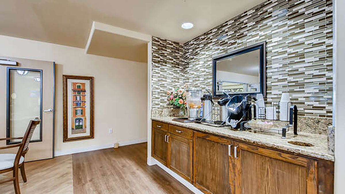 A modern coffee station with wooden cabinets and a stylish tiled backsplash, featuring a mirror and hardwood flooring.