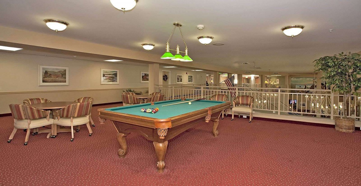 A spacious billiard room with a pool table and green overhead lighting, featuring comfortable seating and decor.