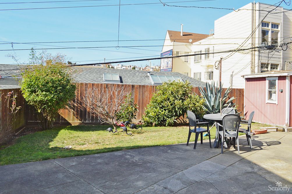 A sunny backyard with a patio area featuring a table and chairs, surrounded by greenery and enclosed by a wooden fence.