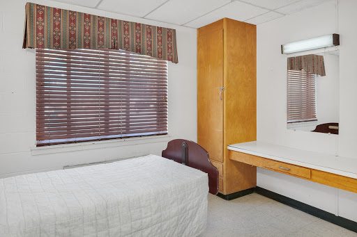 A simple and minimalistic bedroom featuring a bed, wooden furniture, and natural light from the window.