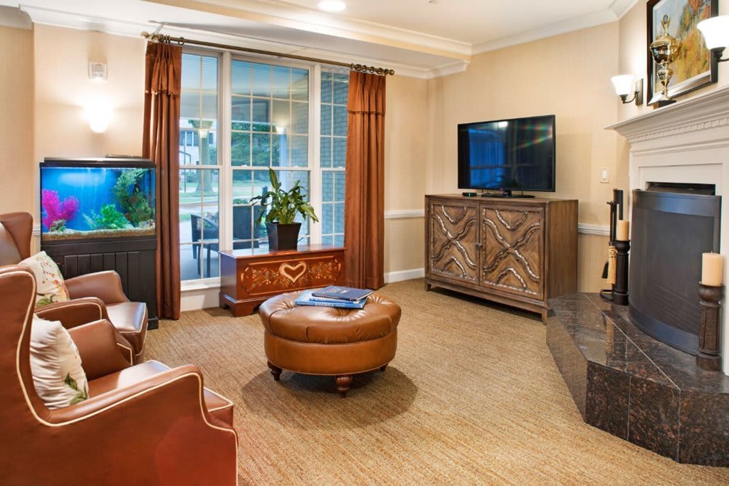 A cozy living room with leather armchairs, an ottoman, a TV on a wooden cabinet, and large windows with curtains.