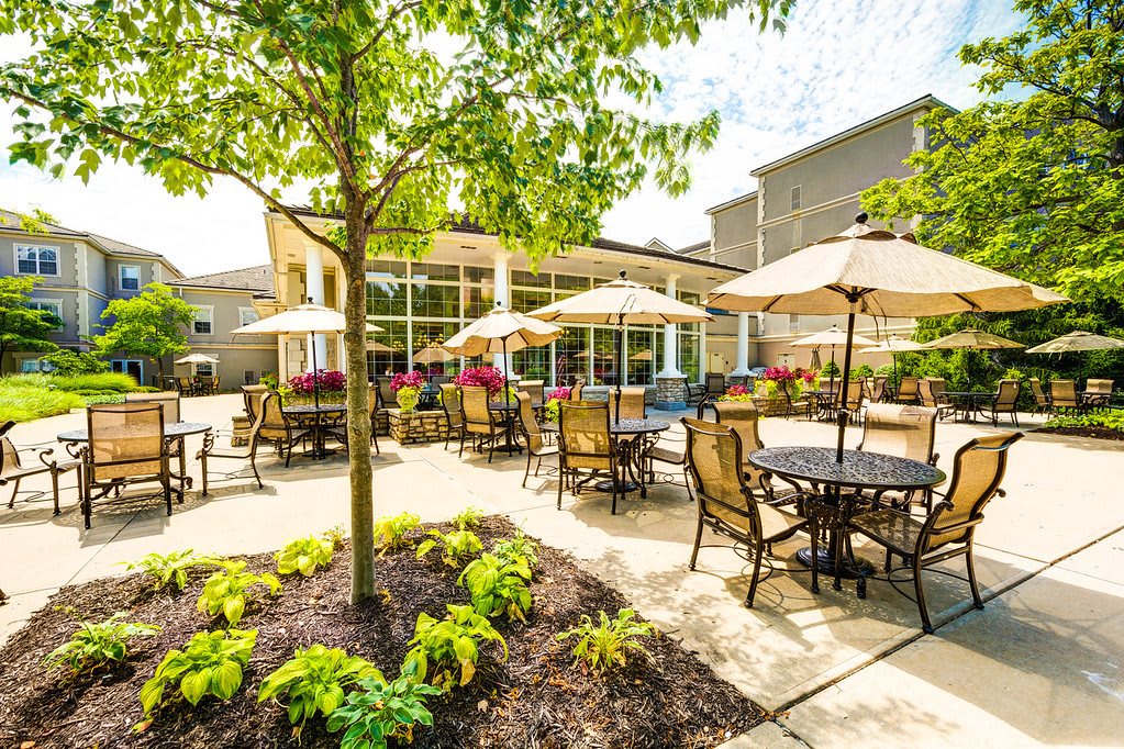 A sunny outdoor patio with tables and chairs shaded by umbrellas, surrounded by lush greenery.