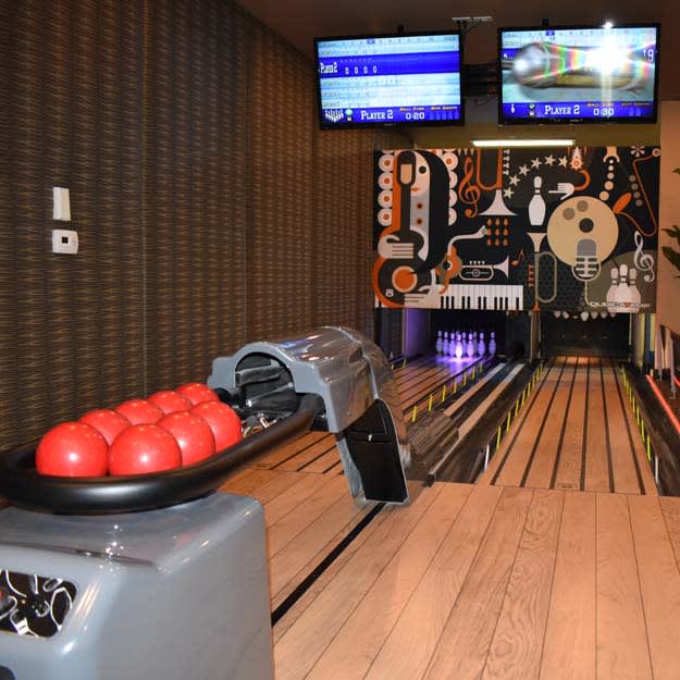 An indoor bowling alley with vibrant red bowling balls and two overhead screens displaying scores.