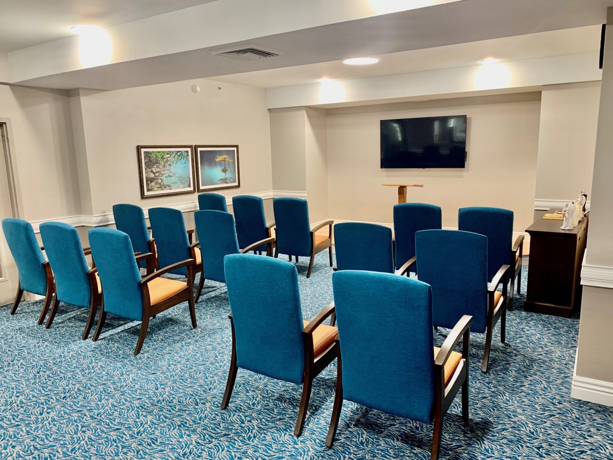 A small conference room with rows of blue upholstered chairs facing a wall-mounted television.