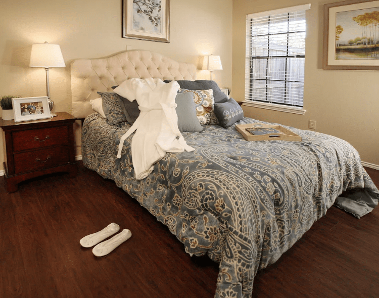 A cozy bedroom with a neatly made bed, featuring patterned bedding and several decorative pillows. A lamp sits on the nightstand beside the bed, and natural light filters through the window.