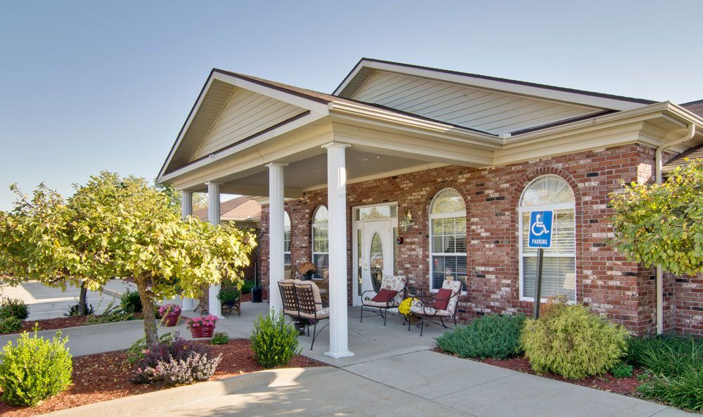 A welcoming building entrance with brick walls, white columns, and outdoor seating surrounded by landscaping.