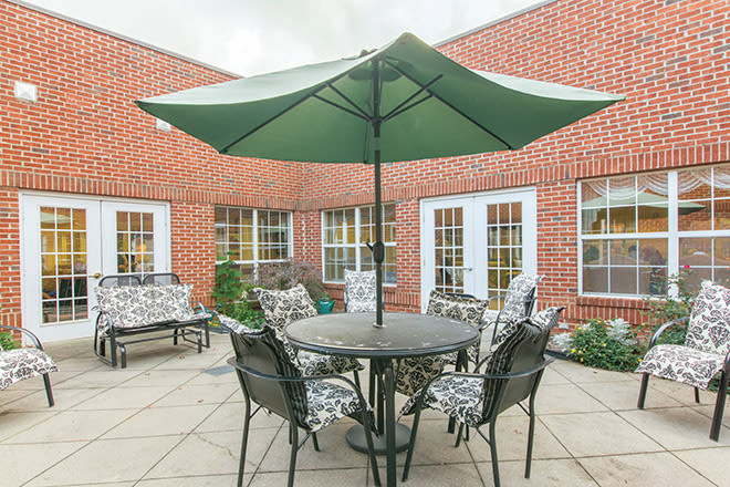 A cozy outdoor patio area featuring a table with chairs and a large green umbrella, surrounded by brick walls and windows.