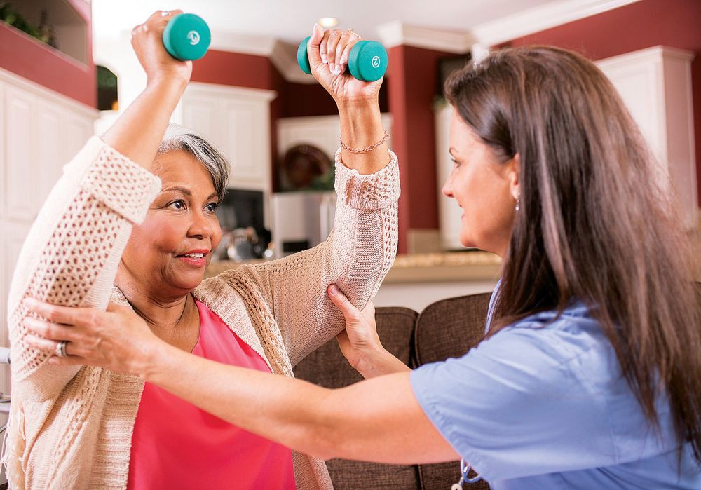 A therapist assists an elderly woman in exercising with dumbbells at home.