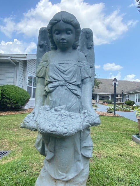 A stone angel statue in a garden setting with buildings in the background under a blue sky.