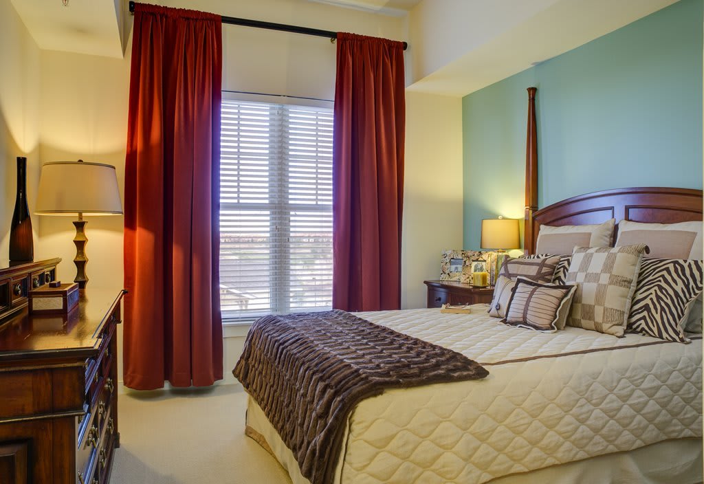 A cozy bedroom with a neatly made bed, adorned with pillows and a blanket, featuring red curtains and soft lighting.