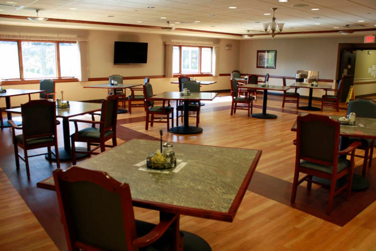 An empty dining room with tables and chairs arranged on a hardwood floor, featuring a television and large windows.