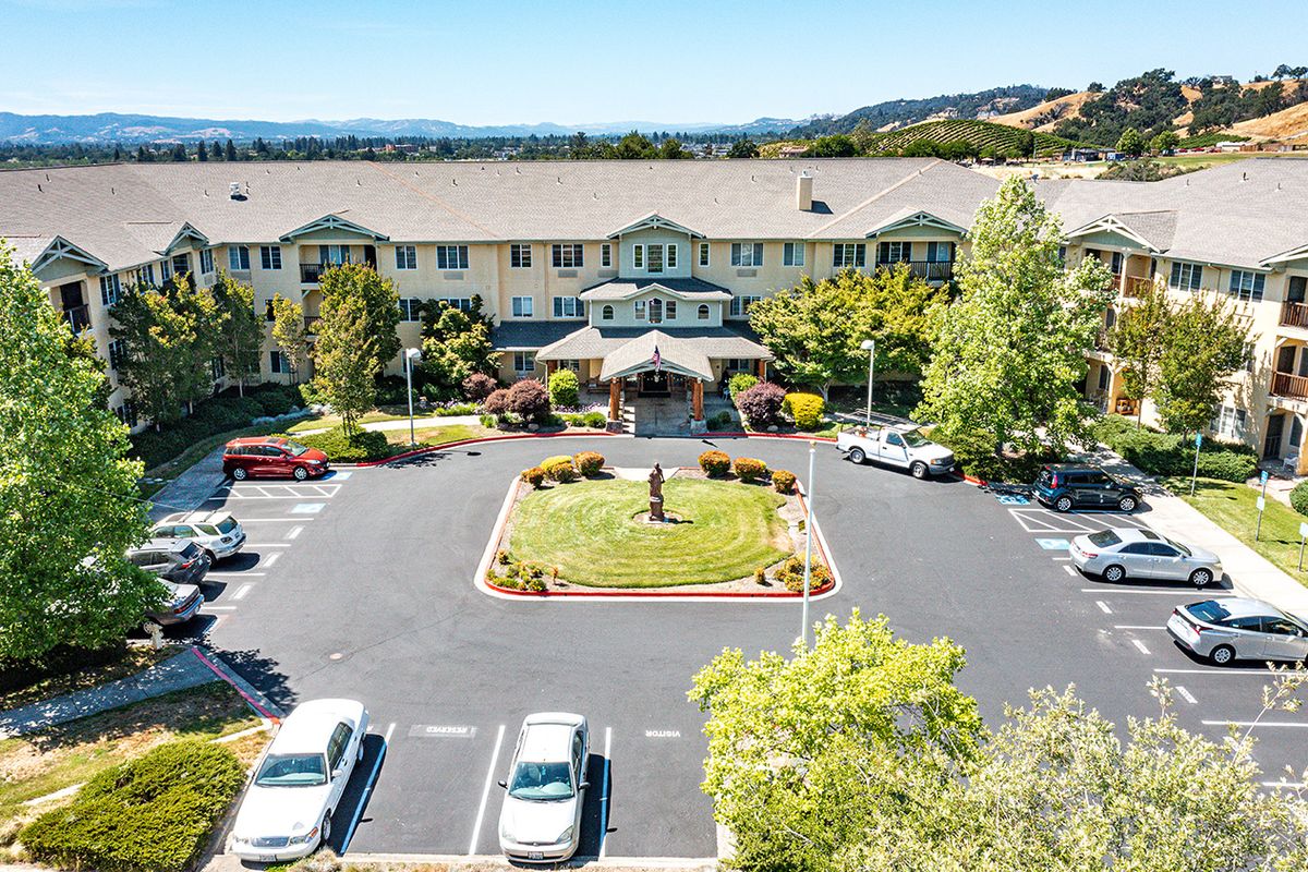 An aerial view of a residential building with a central entrance, surrounded by a parking lot and landscaped greenery on a sunny day.