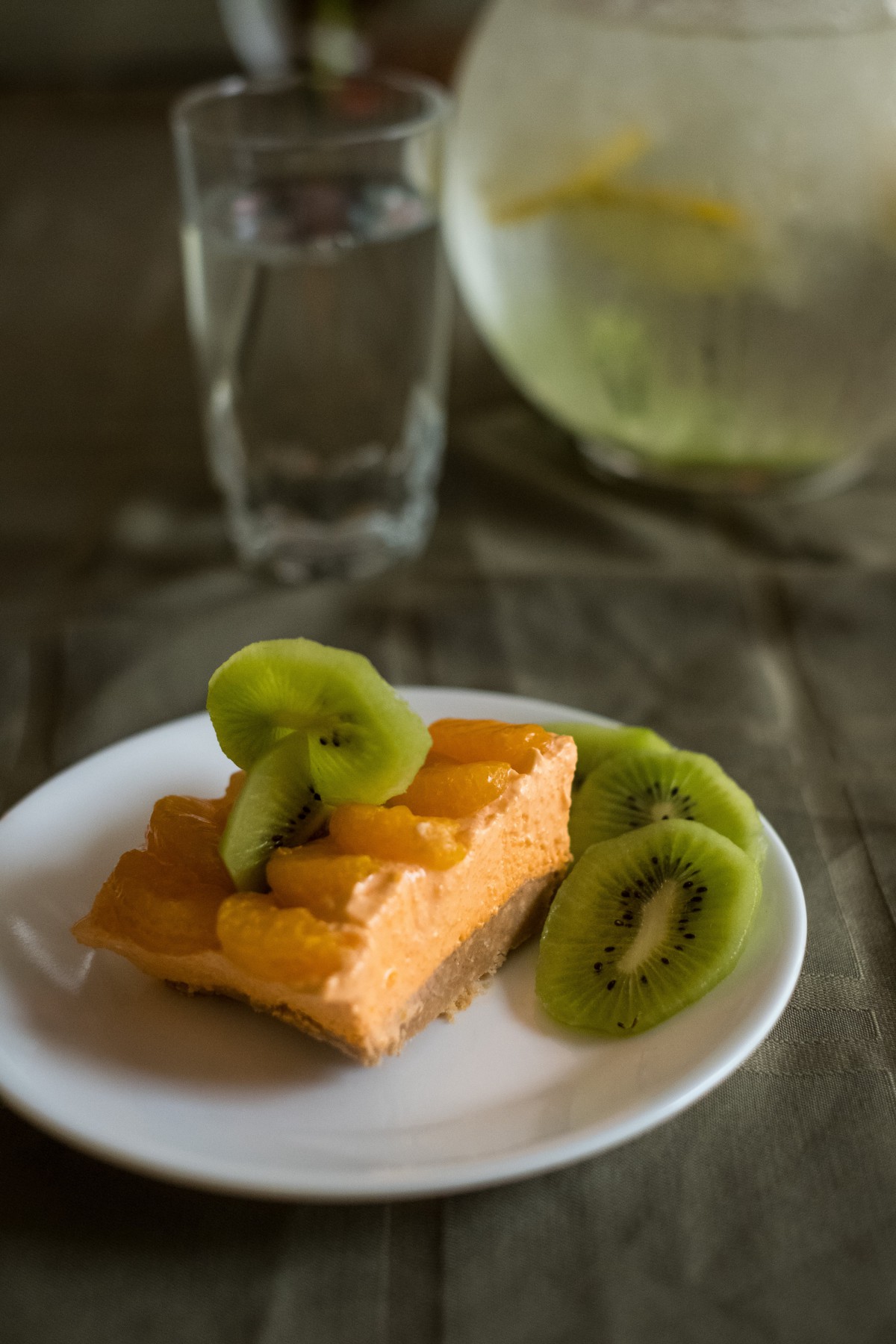 A slice of orange cheesecake topped with kiwi slices on a white plate, accompanied by a glass of water.