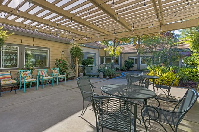 A cozy patio with metal furniture under a wooden pergola, surrounded by lush greenery.