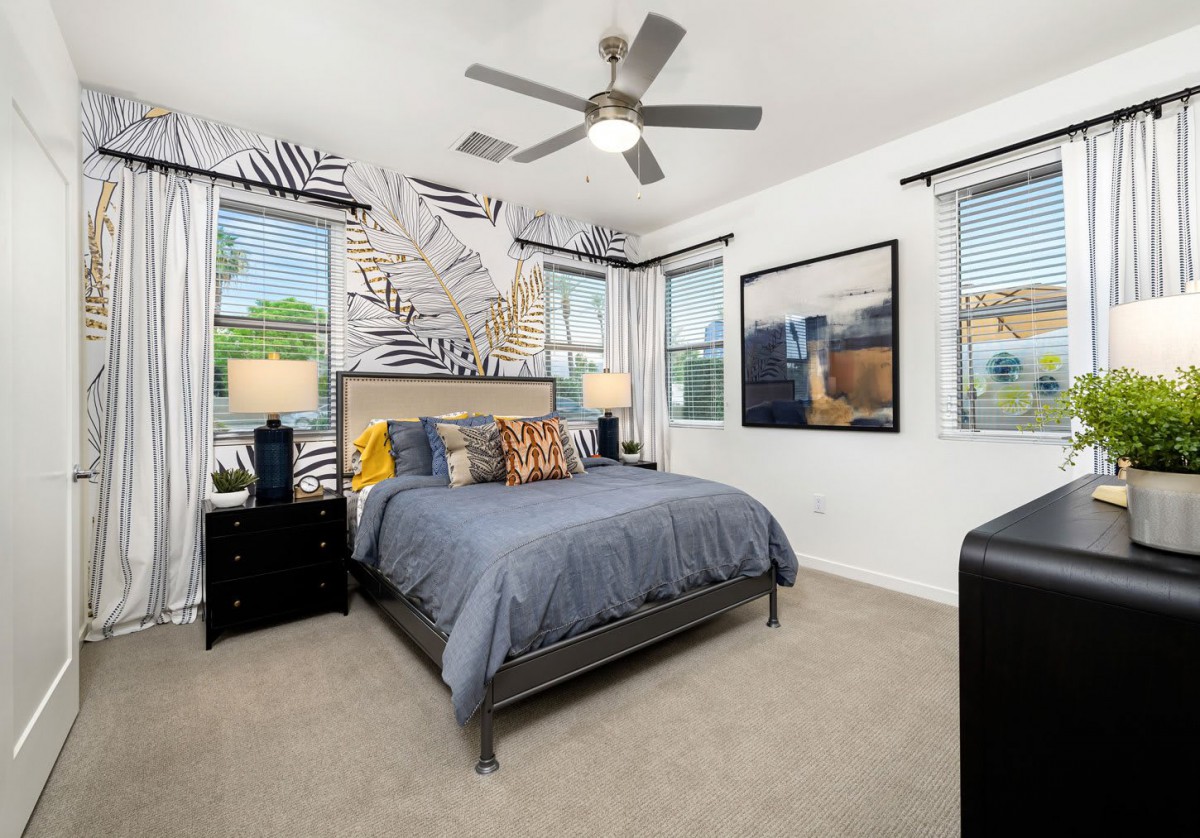 A modern bedroom with stylish decor, featuring a bed with blue bedding, two windows, a ceiling fan, and artistic wall designs.