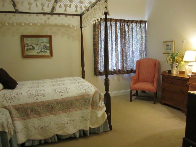 A cozy bedroom featuring a four-poster bed with a canopy and quilt, alongside an armchair, dresser, and window with curtains.