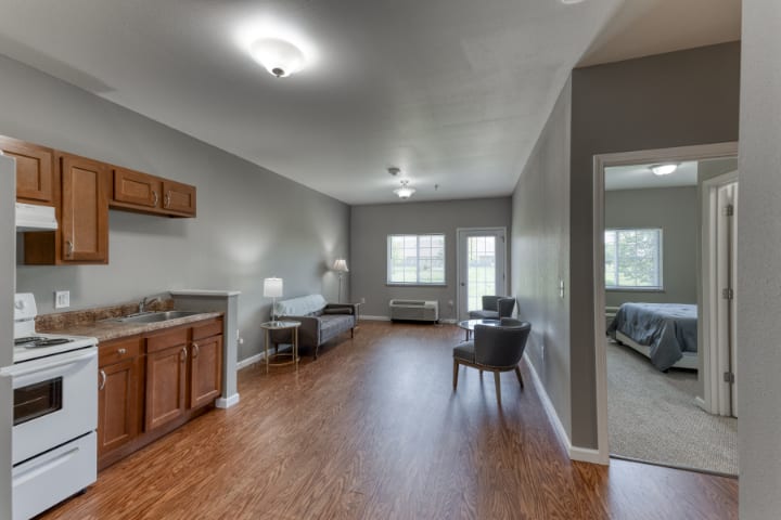 A modern living area with hardwood floors, featuring a sofa and kitchenette, well-lit by natural light from large windows.