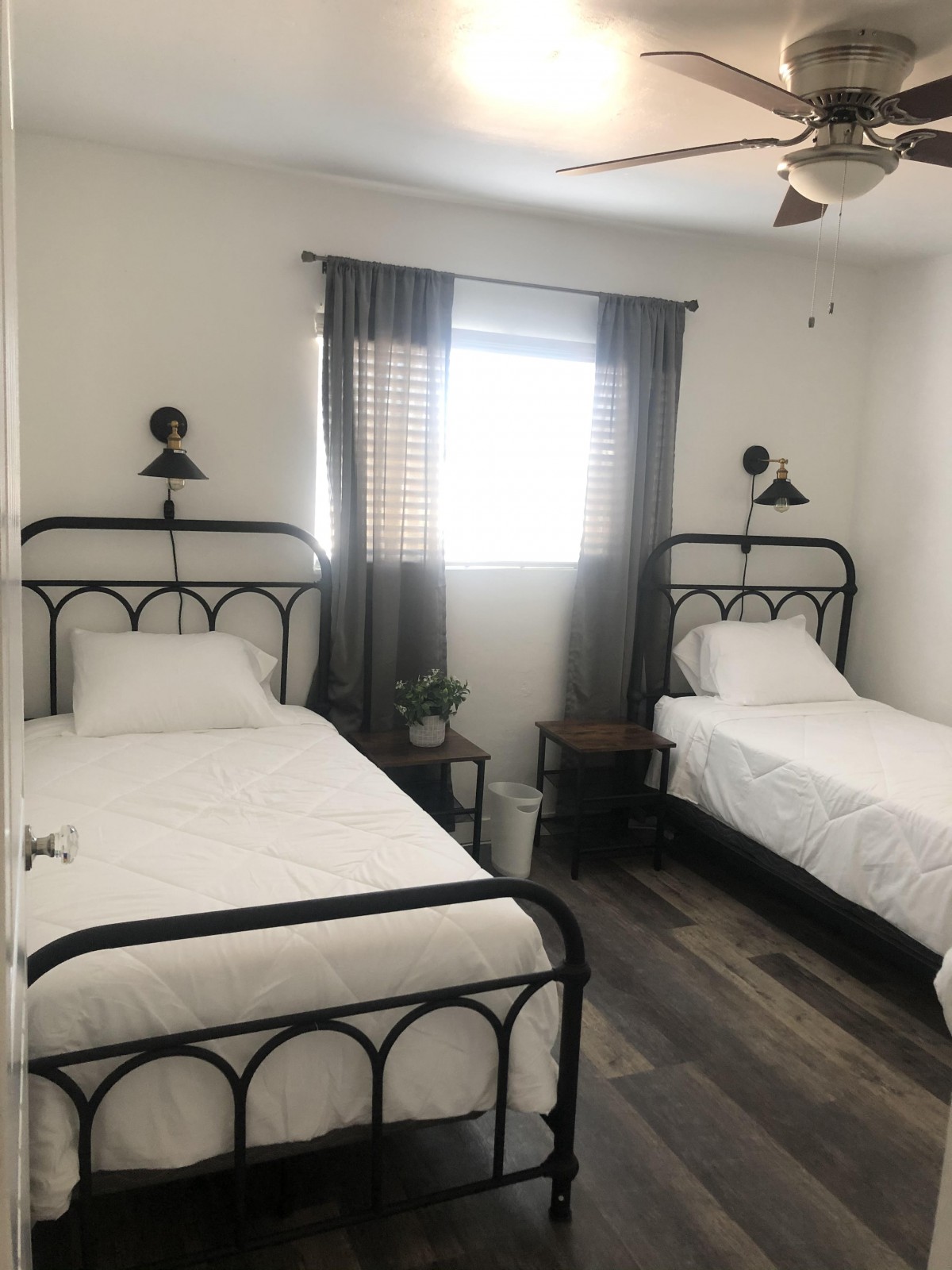 A cozy bedroom featuring two iron-framed beds with white bedding, complemented by wall-mounted lamps and a window with curtains.