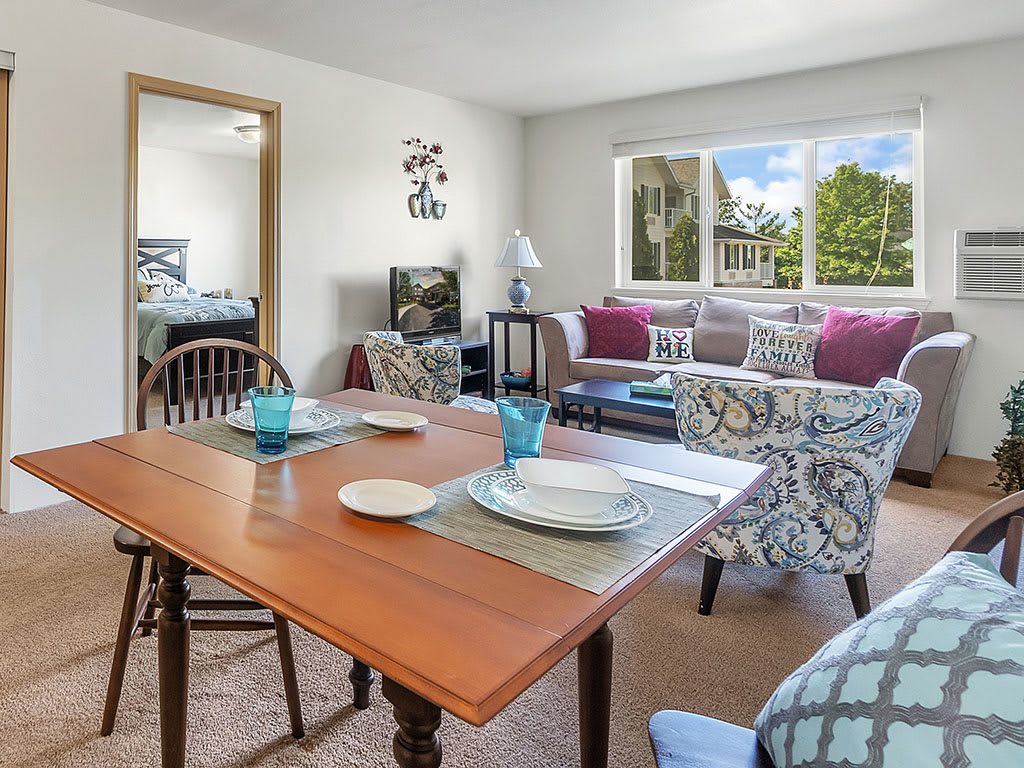 A cozy living and dining area with a wooden table set for two, comfortable seating, and a large window offering an outdoor view.