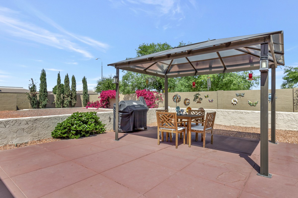 A sunny patio featuring a gazebo with garden furniture and a barbecue grill, surrounded by vibrant bougainvillea and plants.