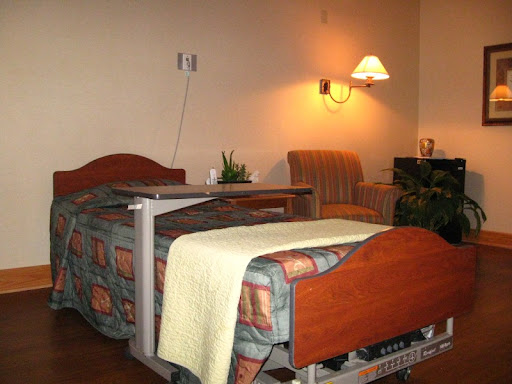 A cozy patient room featuring a bed with a blanket, a chair, and warm lighting.