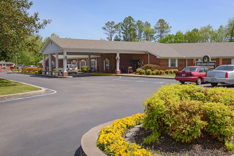 A single-story building with a covered entrance, surrounded by a parking lot and landscaped with flowers and shrubs.