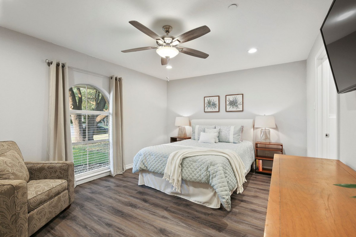 A cozy and modern bedroom with wood flooring, featuring a comfortable bed, nightstands with lamps, a window allowing natural light, a plush chair, and a ceiling fan for ventilation.