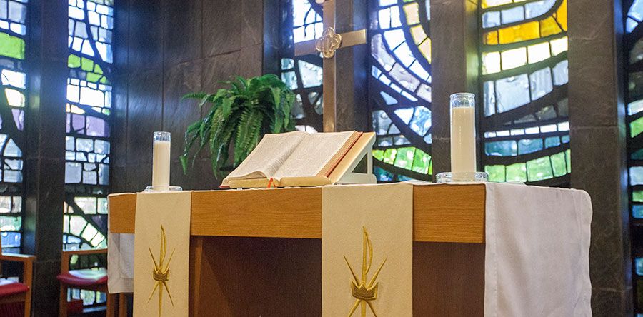A church altar with an open Bible, candles, and a cross against stained glass windows.