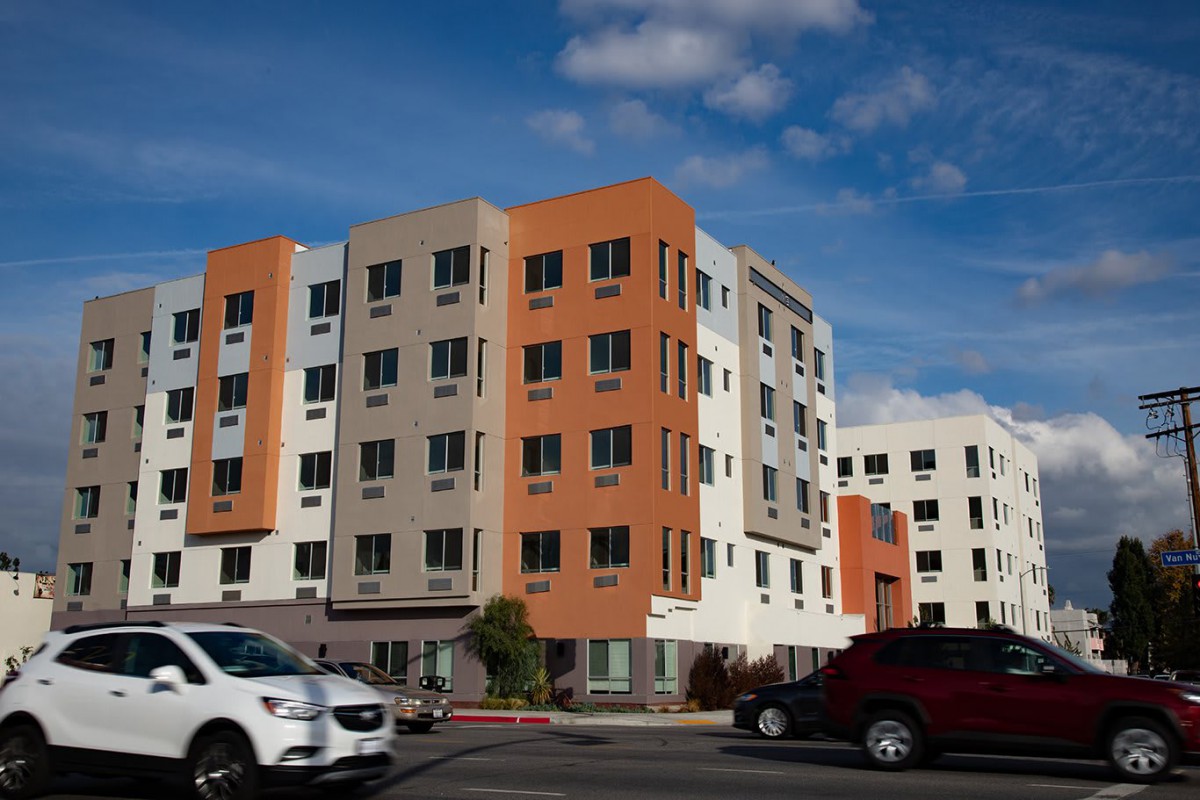 A modern apartment building with orange and white facade, located along a busy street with passing cars.