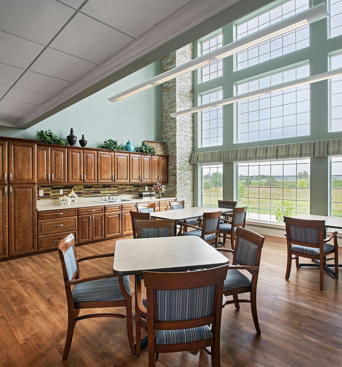 A bright dining room with large windows, wooden cabinets, and several tables and chairs.