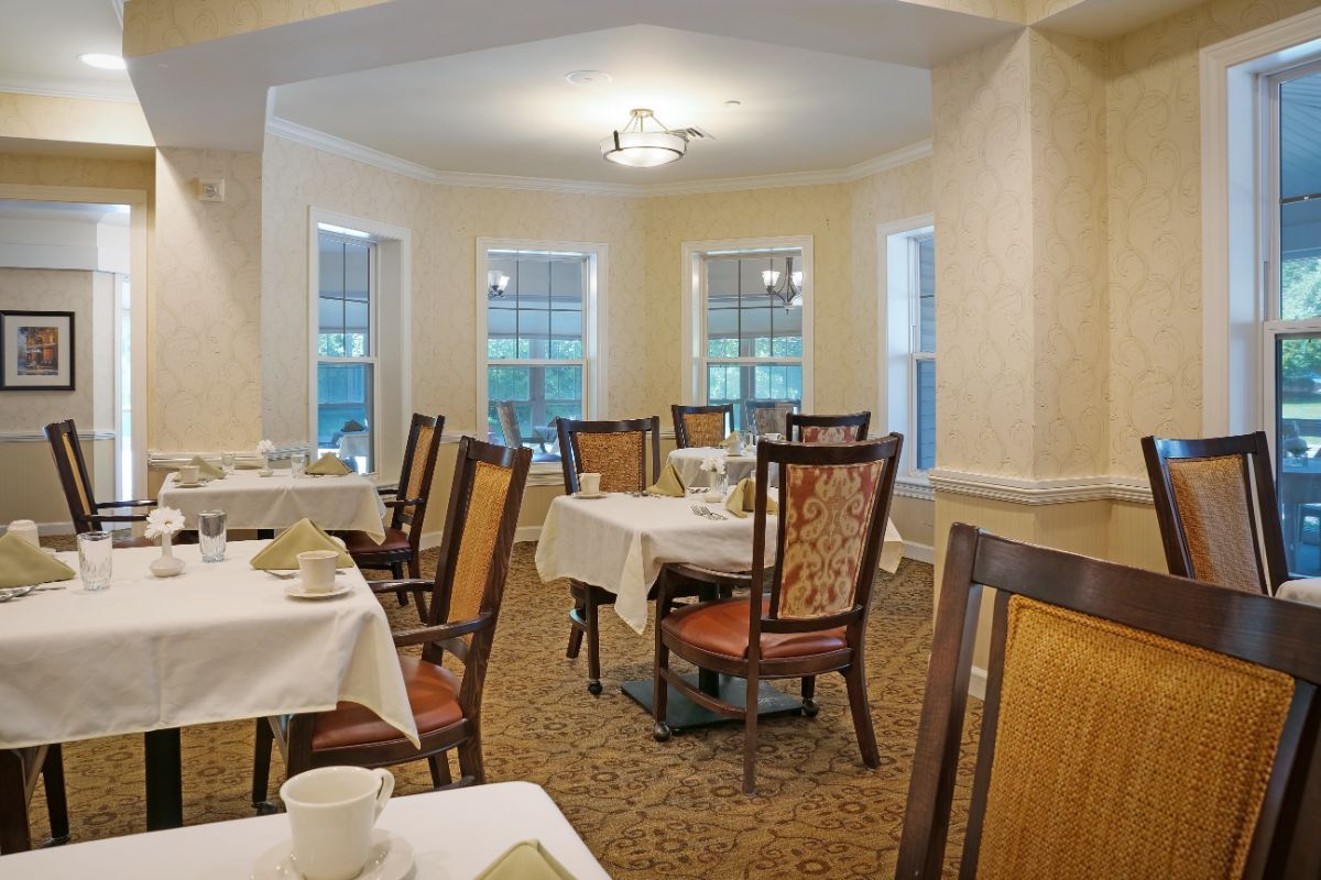 A cozy dining room with neatly arranged tables and chairs, featuring large windows and traditional decor.
