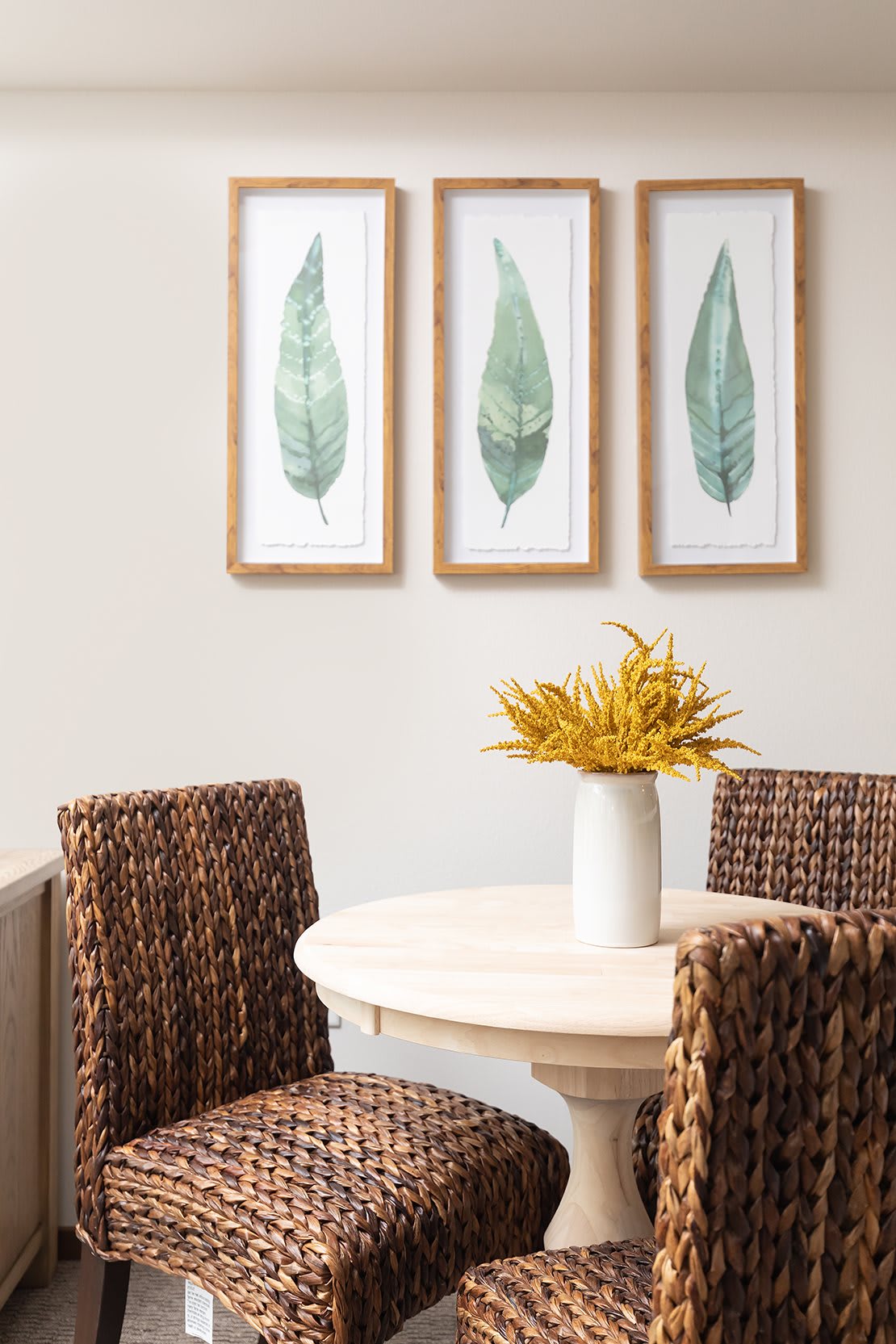 A cozy dining area featuring a round table, wicker chairs, and decorative leaf artwork on the wall.