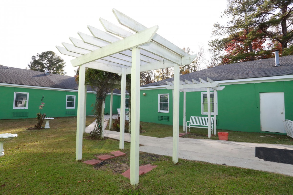 A white pergola stands in a yard with green buildings in the background, featuring a bench nearby.
