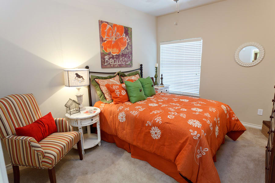 A cozy bedroom with vibrant orange floral bedding and decorative pillows, complemented by wall art and a side table with a lamp.