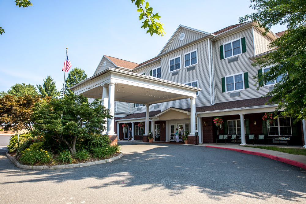 A large residential building with a covered entrance and driveway, featuring a flag and lush landscaping.