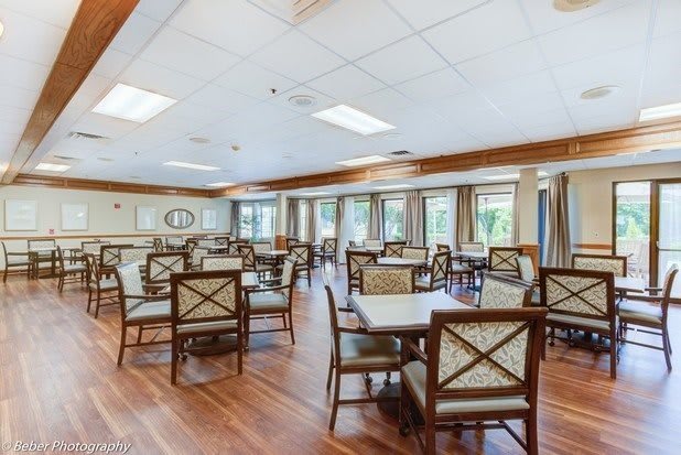 A spacious dining area with multiple tables and chairs, featuring wooden flooring and large windows with curtains.