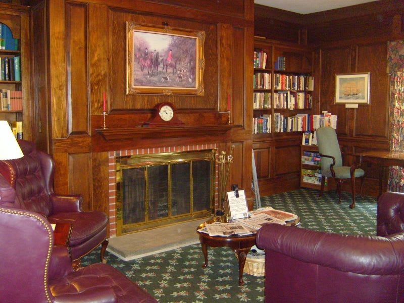 A cozy library room with wood paneling, a fireplace, bookshelves, and comfortable armchairs.