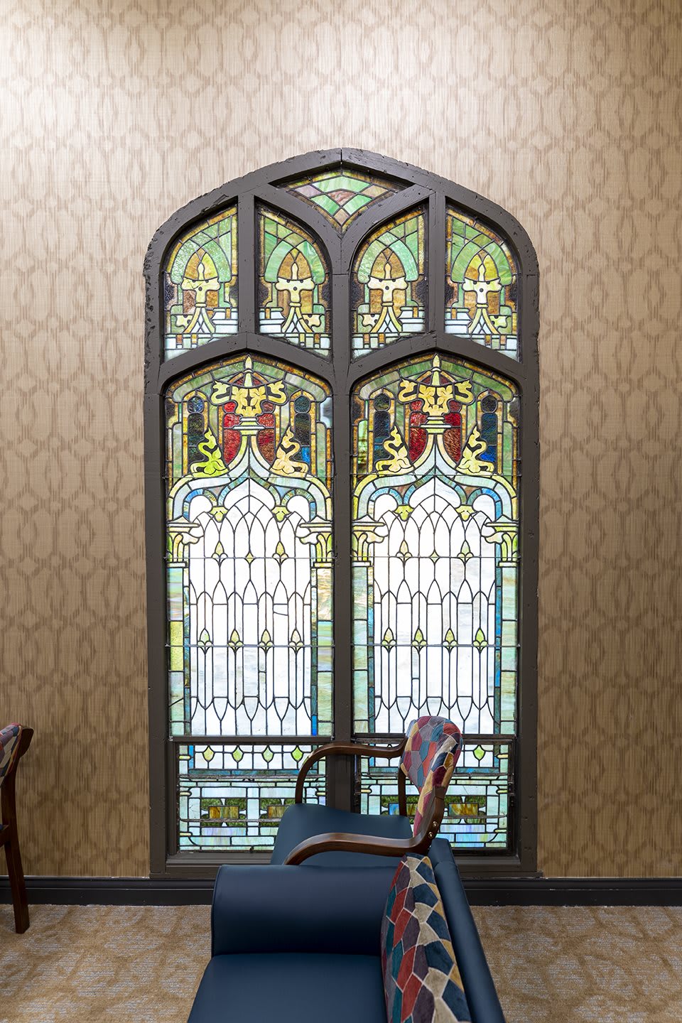 A beautifully designed stained glass window with intricate patterns, framed by an elegant room interior featuring a classic wooden chair.