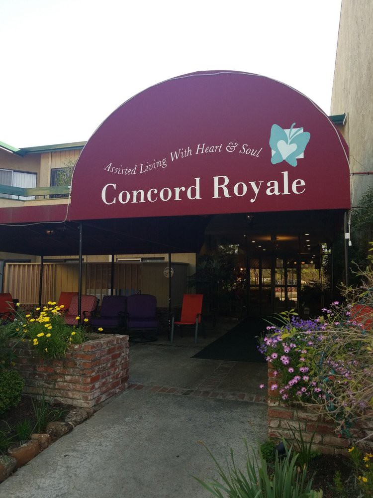 The entrance to Concord Royale is adorned with a maroon awning and surrounded by blooming flowers.