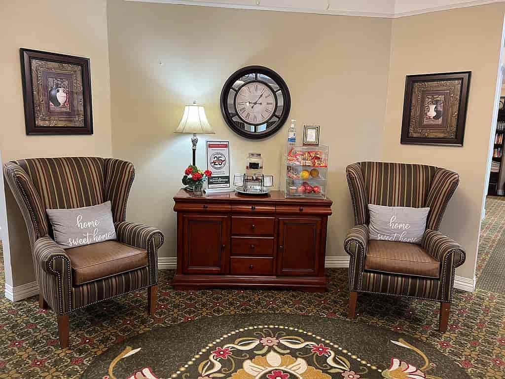 A cozy waiting room with two striped chairs, a central table with a lamp, and a wall clock above.