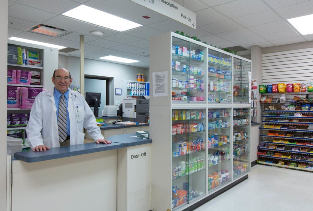 A pharmacist stands behind the counter in a pharmacy, with shelves stocked with various medications.